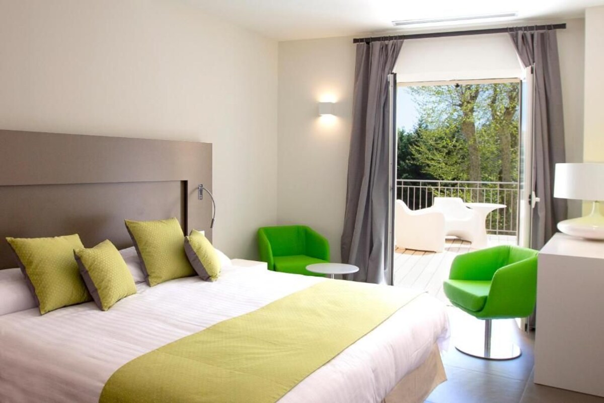 Ein Hotelzimmer mit Bett, grünen Stühlen, einem kleinen Tisch und einem Balkon mit Blick auf Bäume.
