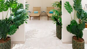 Een hal met een witte vloer, twee rieten stoelen met blauwe kussens, en grote groene planten in potten met een patroon.