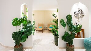 Een modern interieur met een hal die uitkomt in een zithoek, met grote potplanten en een decoratieve metalen boomsculptuur.