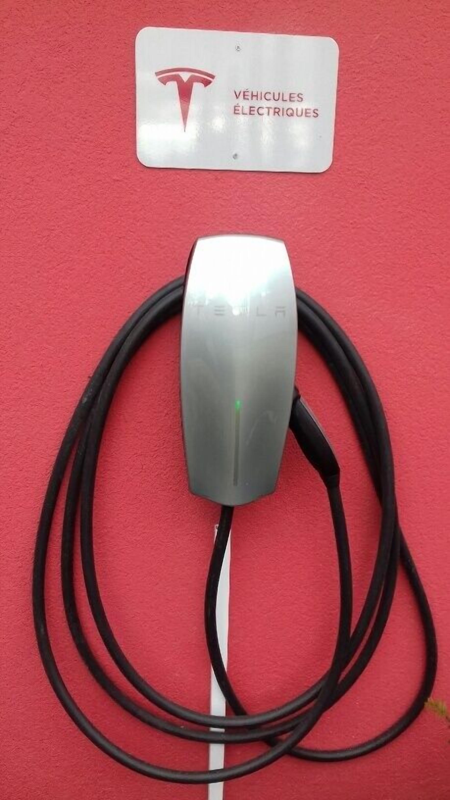 Una estación de carga de Tesla con un conector metálico y un cable, sobre un fondo rojo.