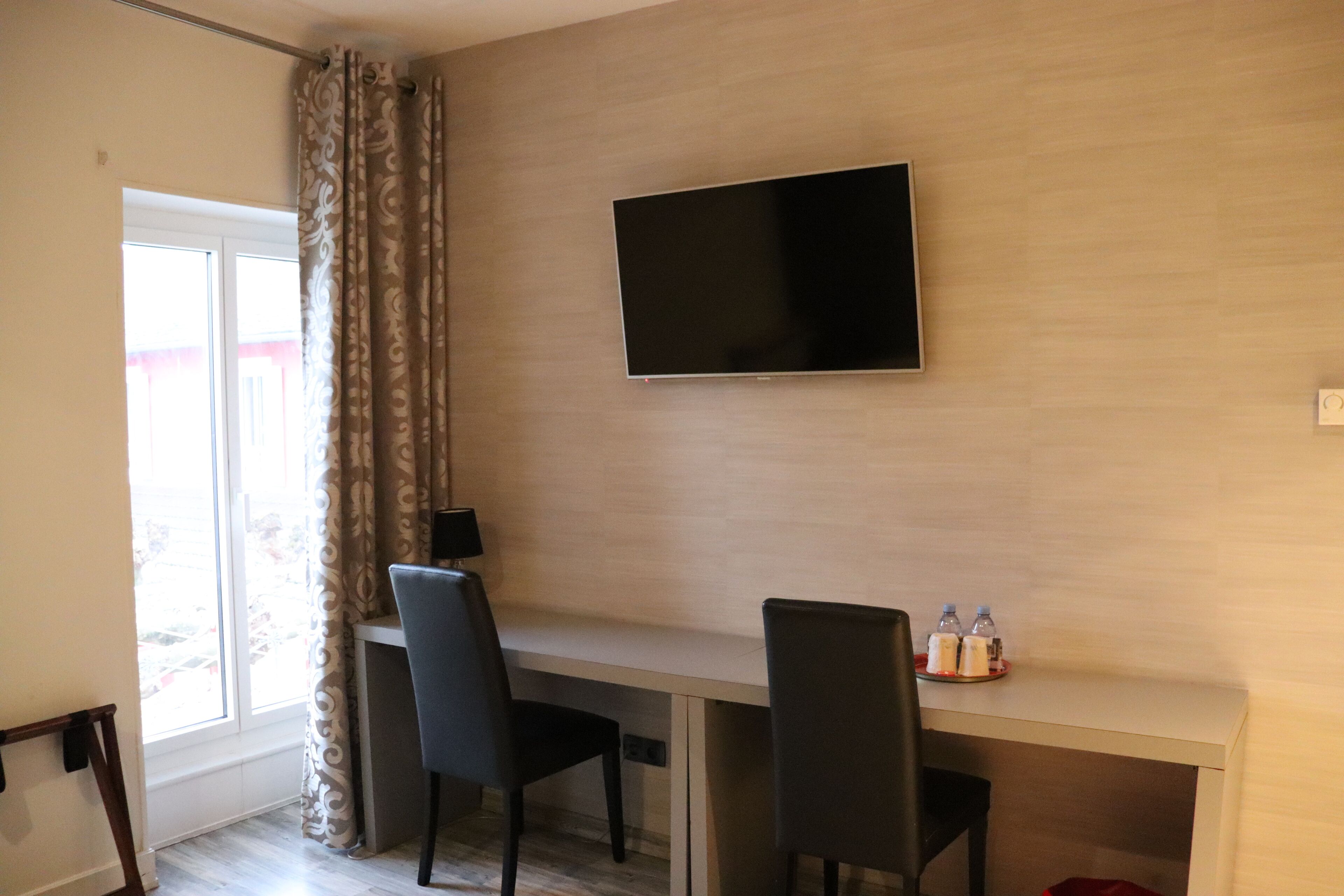 A modern room with a flat-screen TV mounted on the wall, two black chairs, a small table with bottles, and a window with curtains.