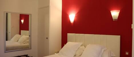 Una habitación de hotel con cama, mesita de noche, espejo y paredes con detalles en rojo.