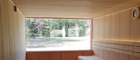 A sauna room with a bench, a wooden cabinet, and a window offering a view of a garden.