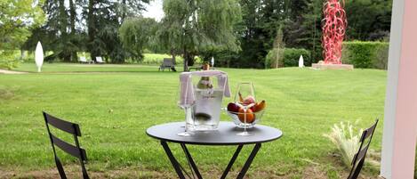 An outdoor seating area with a table set for two, surrounded by a garden with trees and a sculpture.