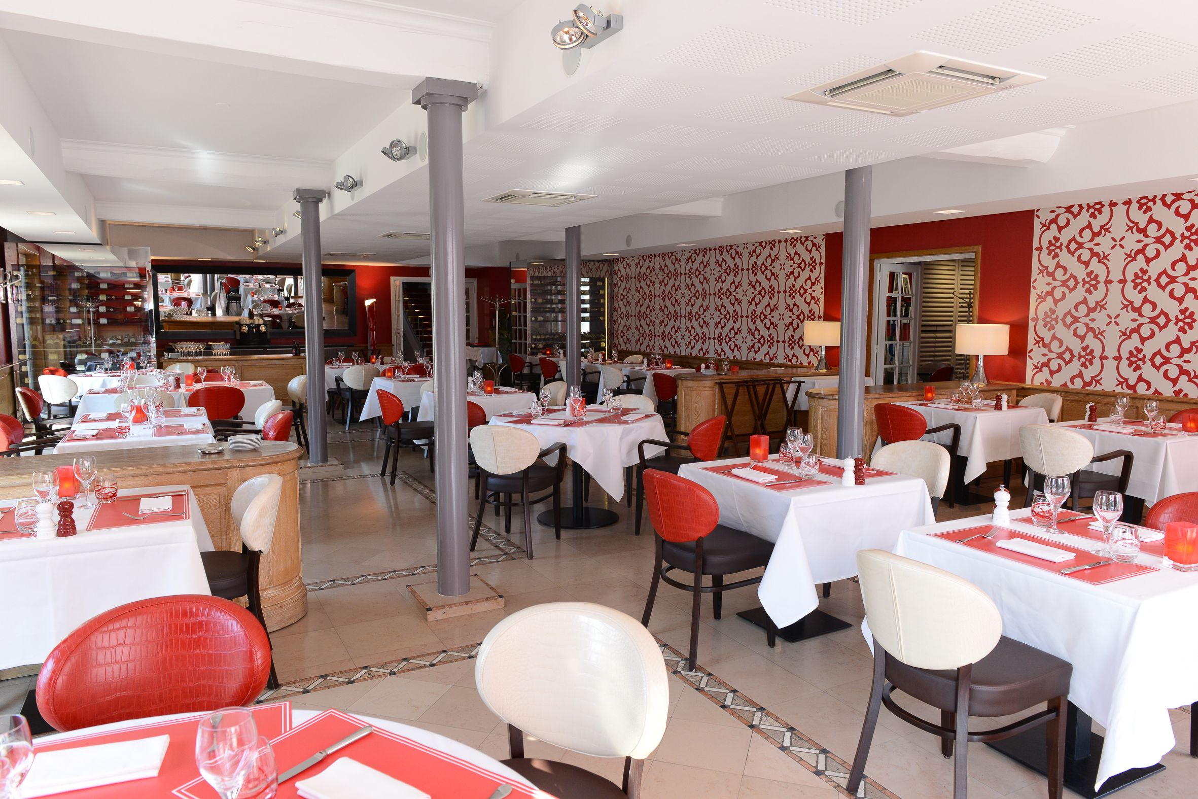 A restaurant with white tables and red chairs, featuring a patterned wallpaper and a bar area in the background.