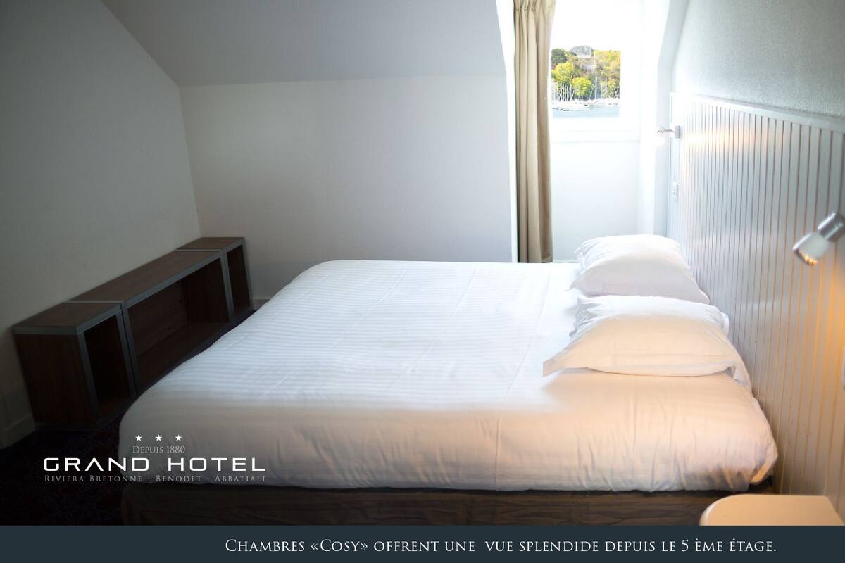 A hotel room with a bed, a bedside table, and a window with curtains. The room has a modern design with white walls and a wooden floor.
