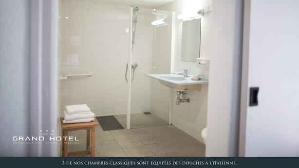 Um banheiro moderno com chuveiro, pia e vaso sanitário. Há um porta-toalhas e um banquinho de madeira. As paredes e o chão são revestidos com azulejos.