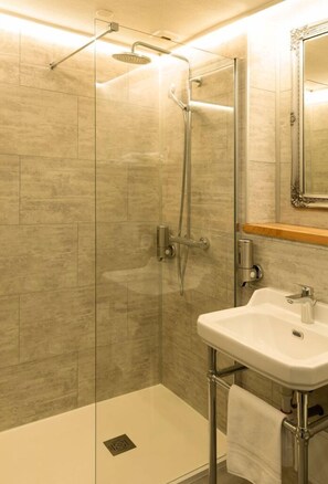 Uma casa de banho moderna com chuveiro em cabine de vidro, lavatório branco e prateleira de madeira sobre o espelho.