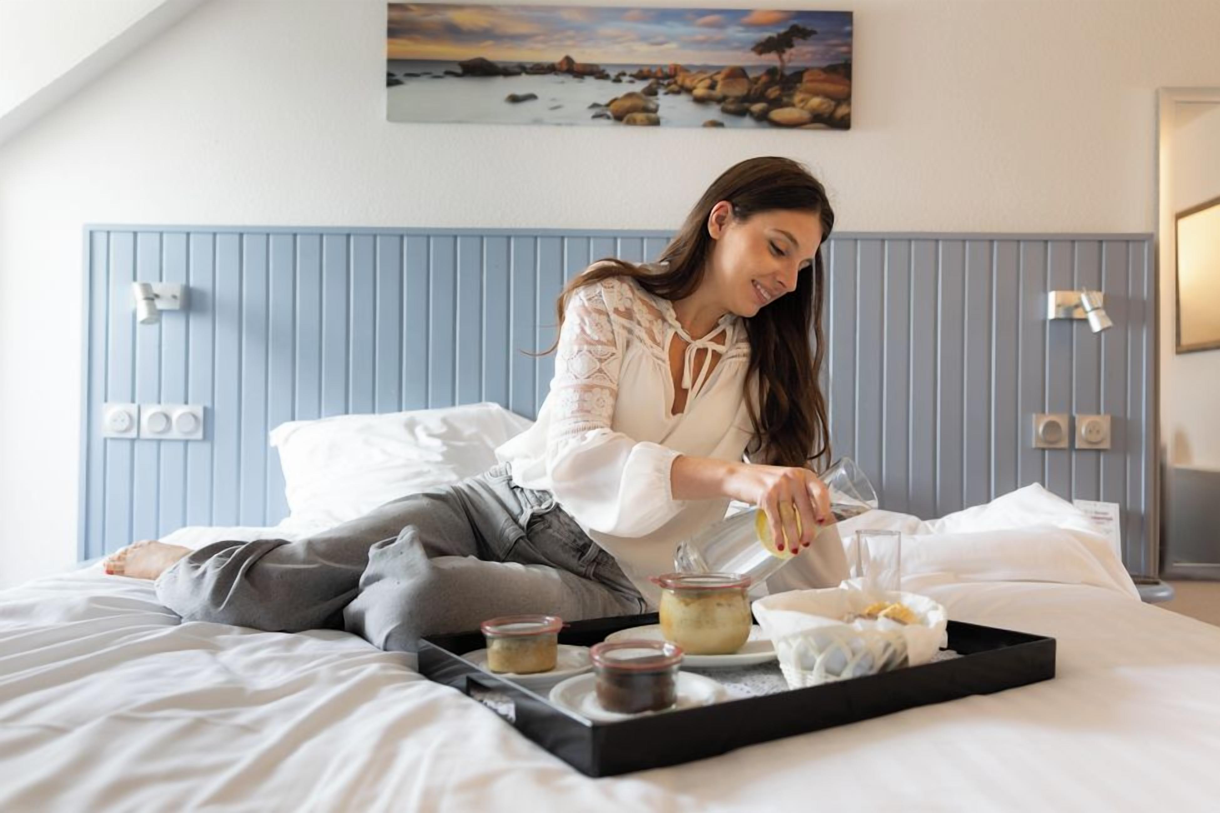 Una persona sentada en una cama con una bandeja que contiene comida, un almohadón y una manta.