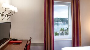 Une chambre d’hôtel avec un lit, un bureau doté d’un ordinateur et d’un classeur rouge, une fenêtre avec des rideaux et une vue sur un port de plaisance.