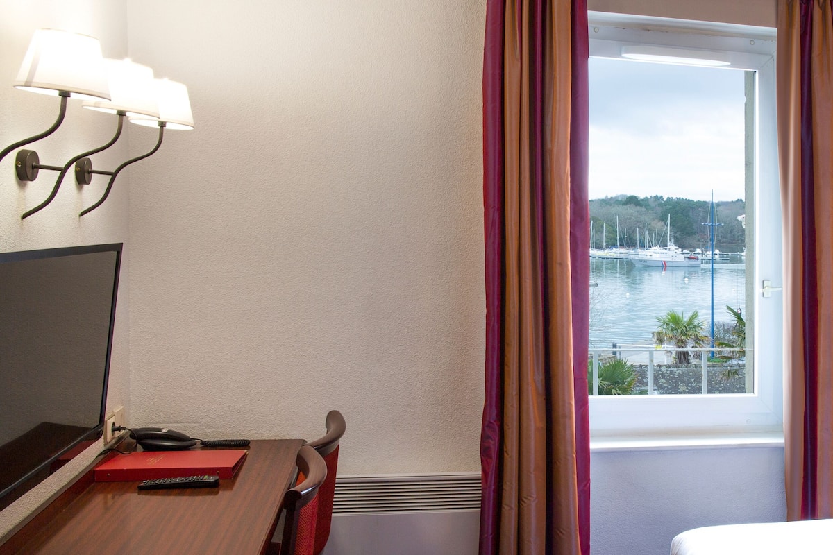 A hotel room with a bed, a desk with a computer and a red folder, a window with curtains, and a view of a marina.