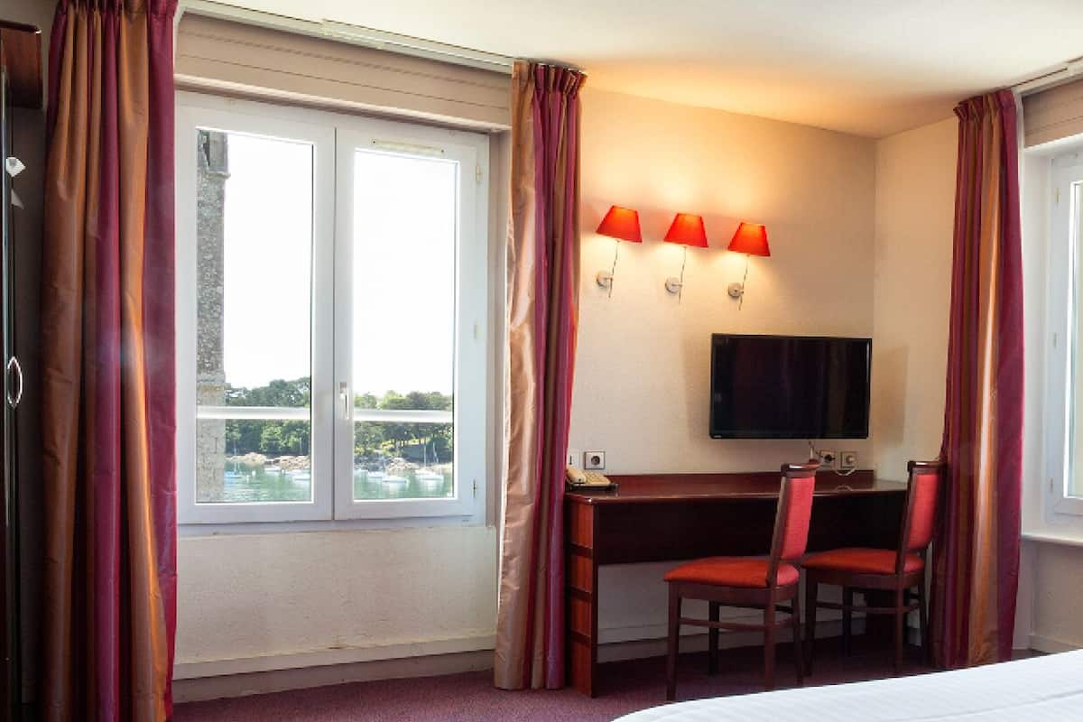 A hotel room with a bed, a desk with two chairs, a television, and a window with curtains.