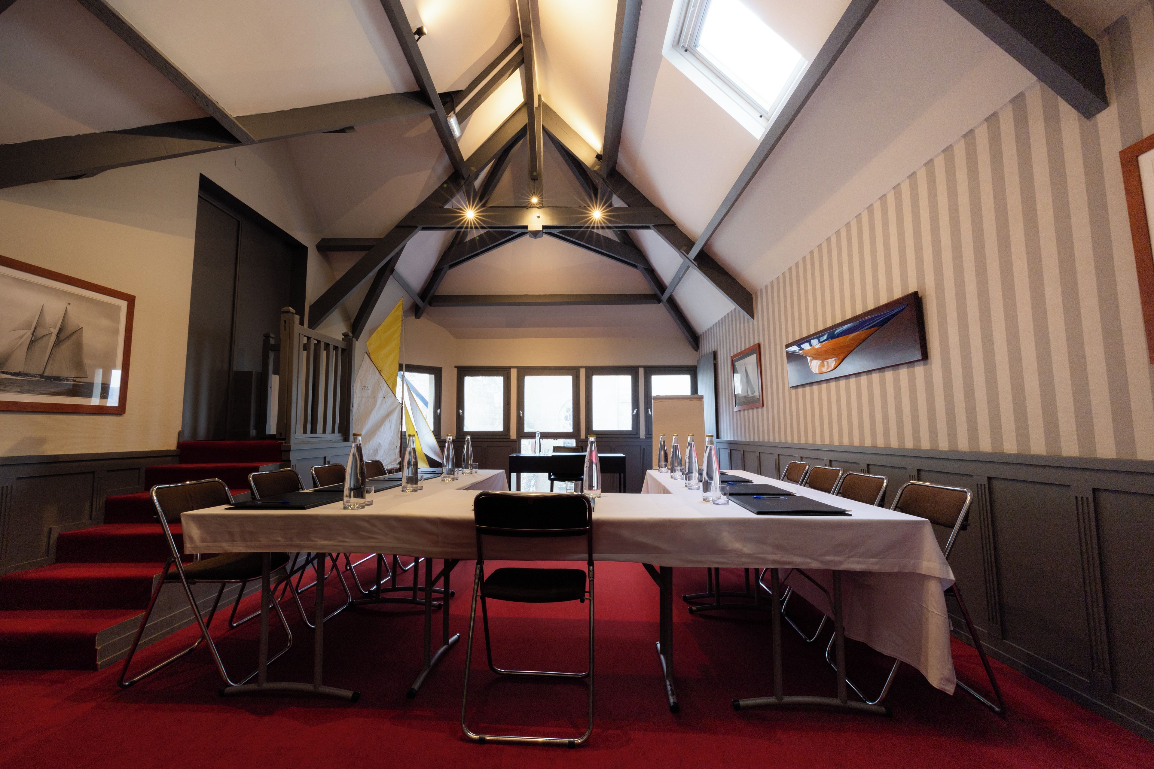 Een vergaderzaal met een lange tafel, stoelen en een rode vloerbedekking.