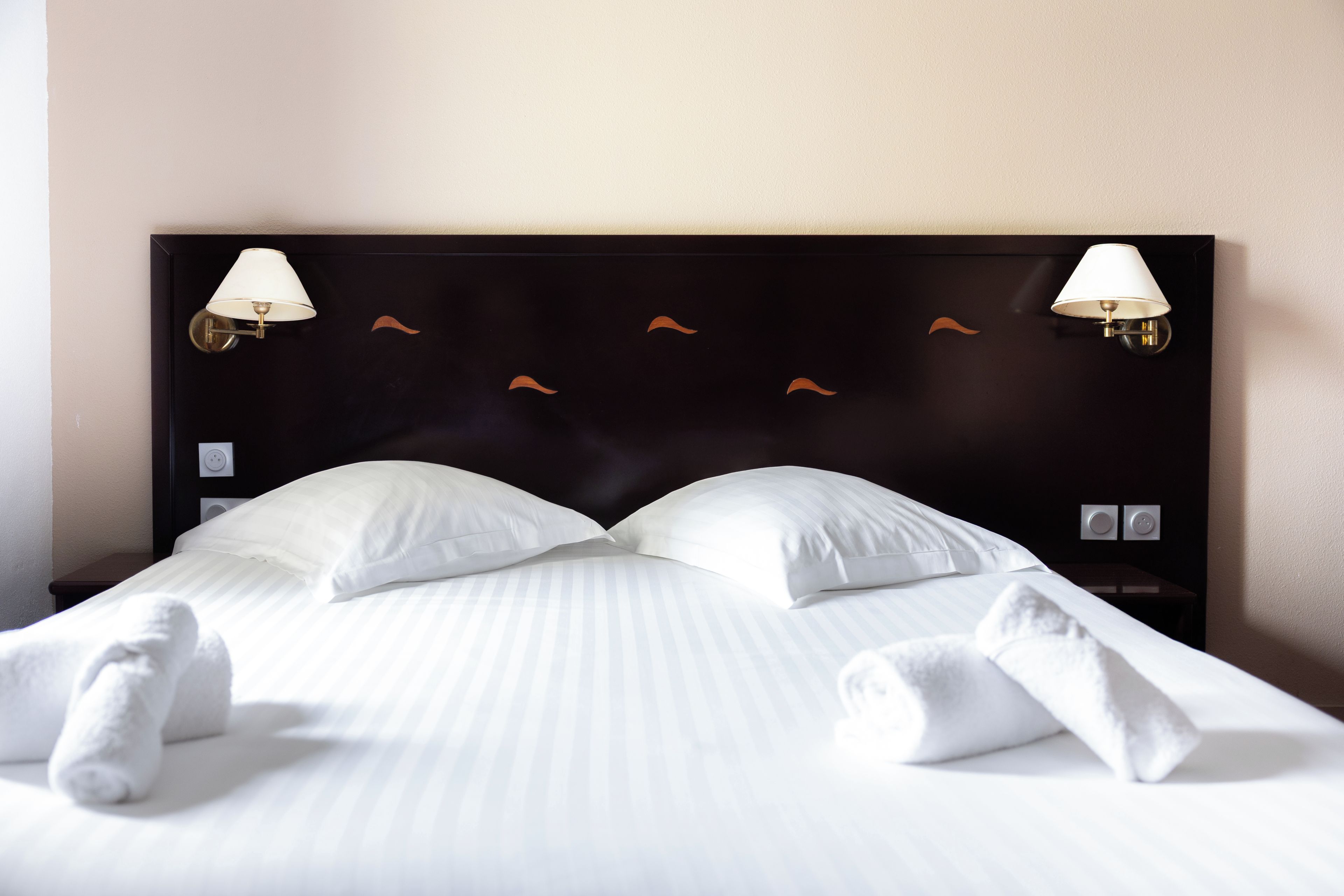 A neatly made bed with white linens and towels, a dark headboard with two wall-mounted lamps, and a simple wall design.