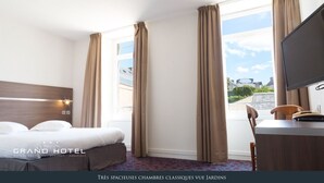 Quarto de hotel com uma cama grande, uma escrivaninha e uma televisão. O quarto possui janelas amplas com cortinas e vista para o exterior.
