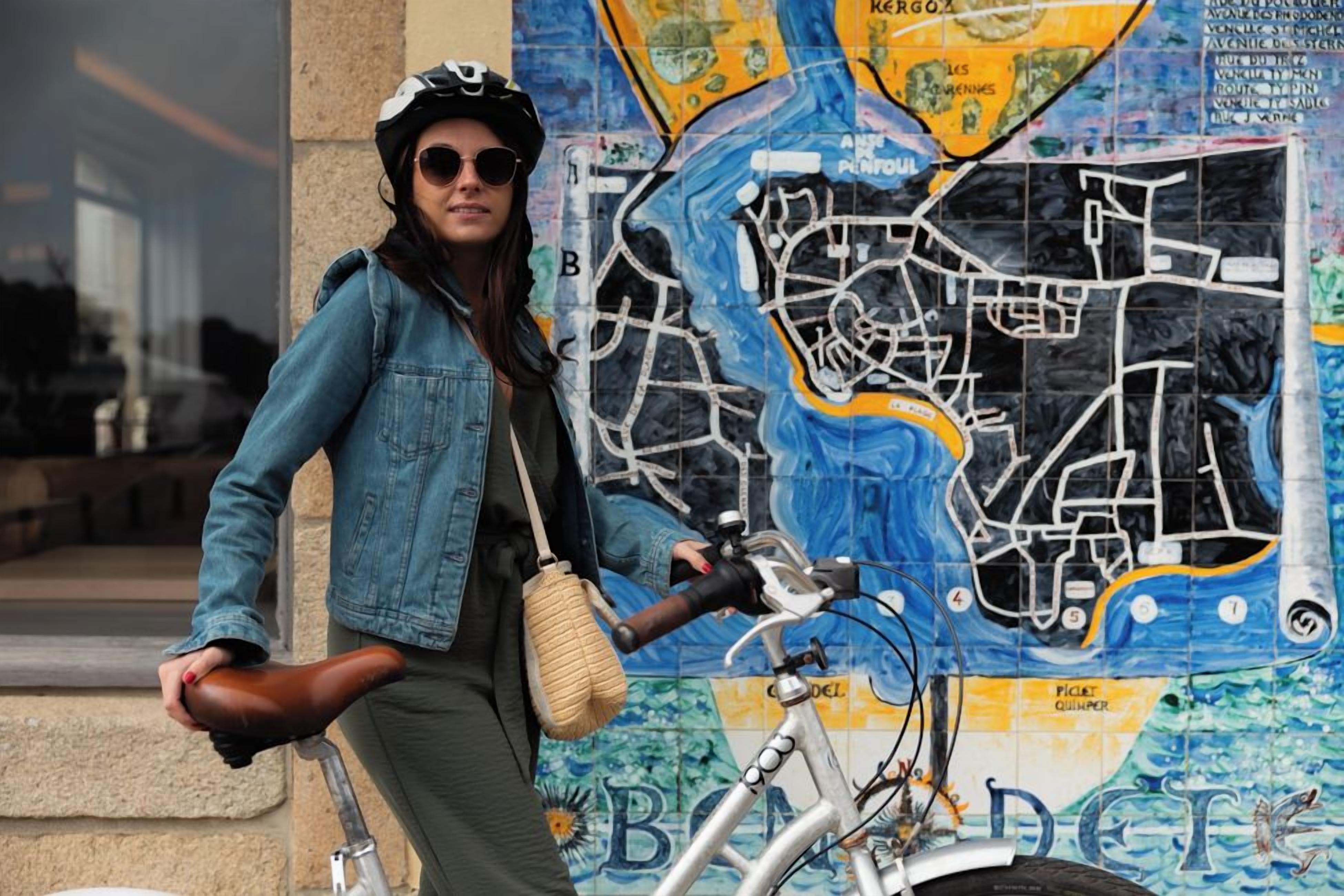 Une personne se tient à côté d’un vélo, avec une fresque représentant une carte en arrière-plan.