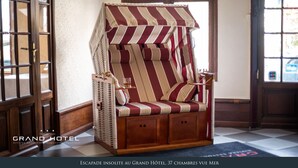 Un fauteuil à rayures, doté d’un dossier tissé et d’un piétement en bois, est placé dans un coin d’une pièce dont le sol est à carreaux et où se trouve une porte sur le côté gauche.