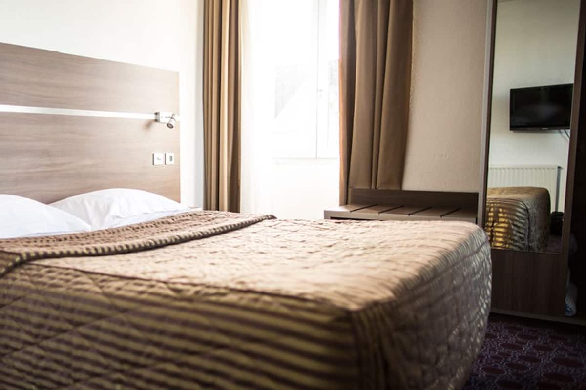 A hotel room with a wooden headboard, a bed with a patterned blanket, a bedside table, and a television mounted on the wall.