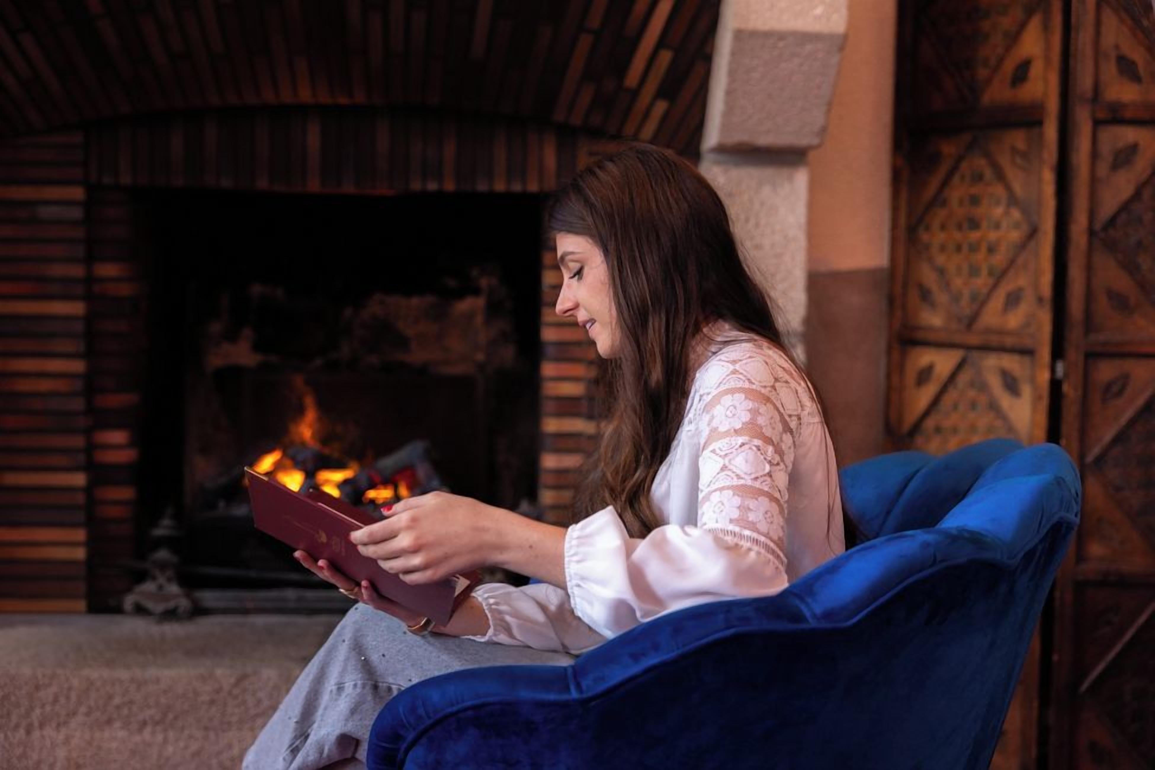 Una persona sentada en una silla azul leyendo un libro, con una chimenea al fondo.