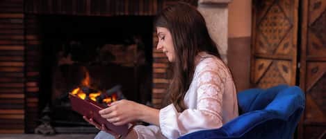 Una persona sentada en una silla azul leyendo un libro, con una chimenea al fondo.