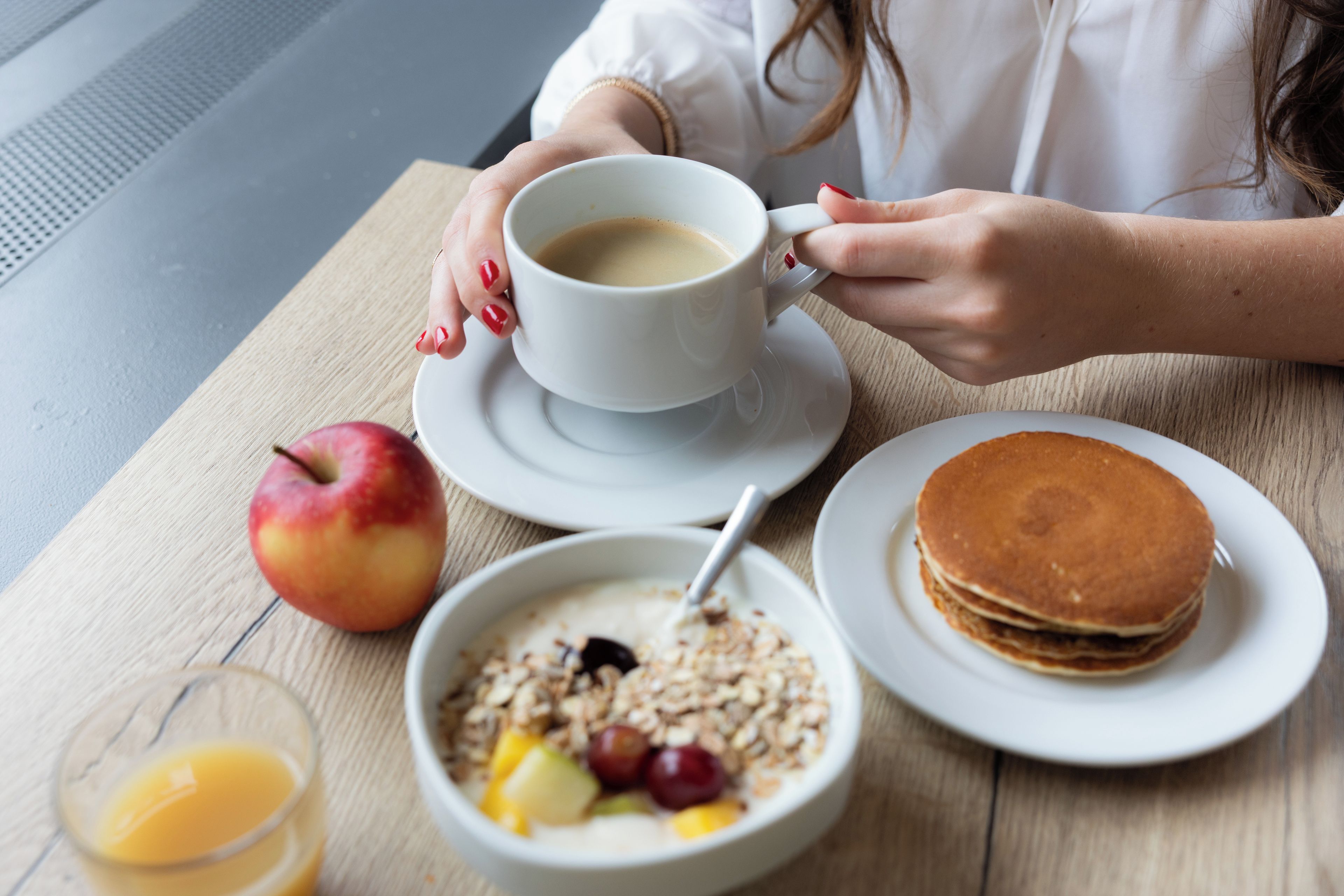 A breakfast setting with a cup of coffee, a plate of pancakes, a bowl of cereal with fruits, and an apple on a wooden table.