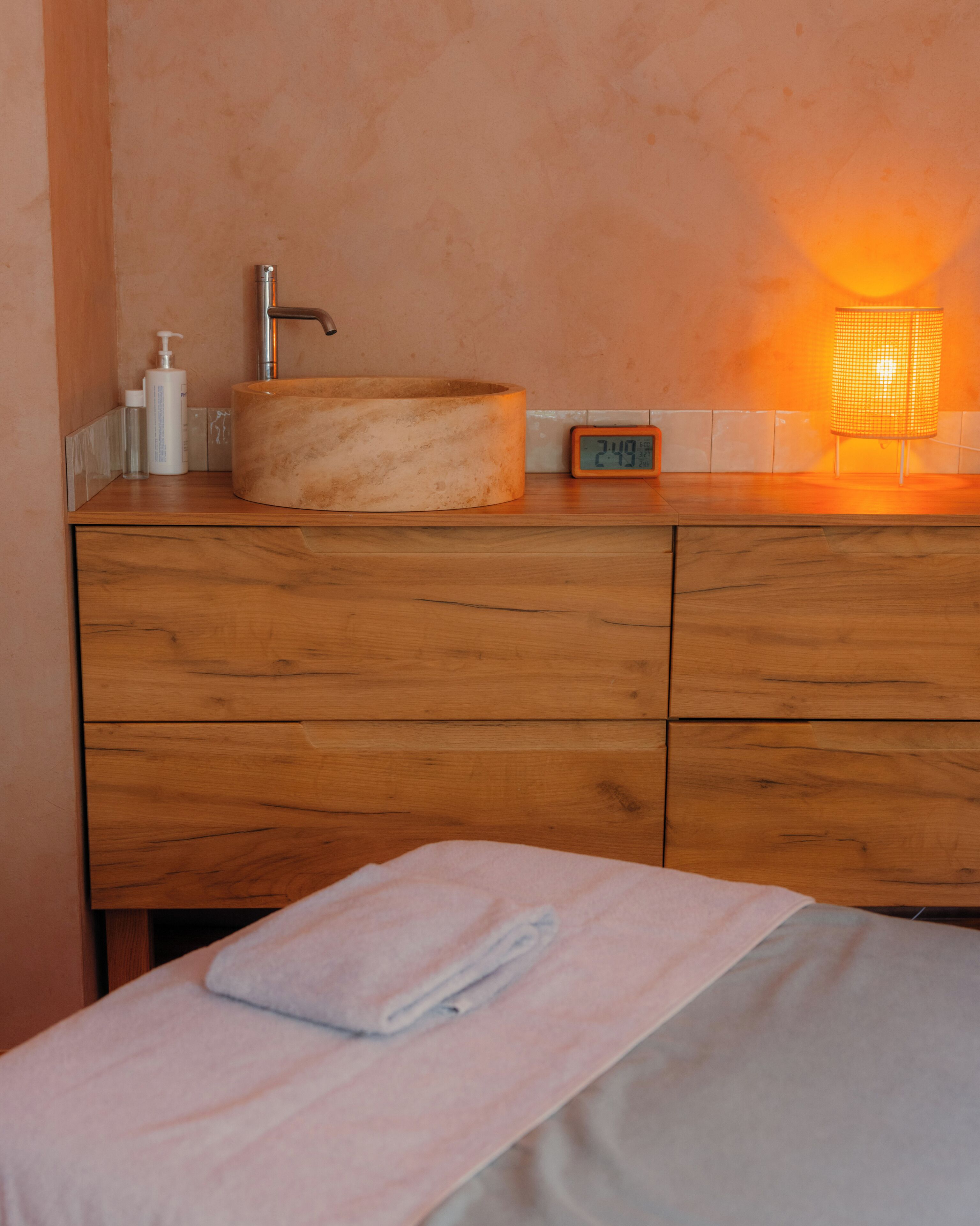 Une salle de bain avec un meuble-lavabo en bois, un lavabo rond, un robinet, une bouteille de shampoing, un verre d’eau, une horloge et une lampe allumée.