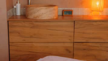 A bathroom with a wooden vanity, a round sink, a faucet, a bottle of shampoo, a glass of water, a clock, and a lit lamp.