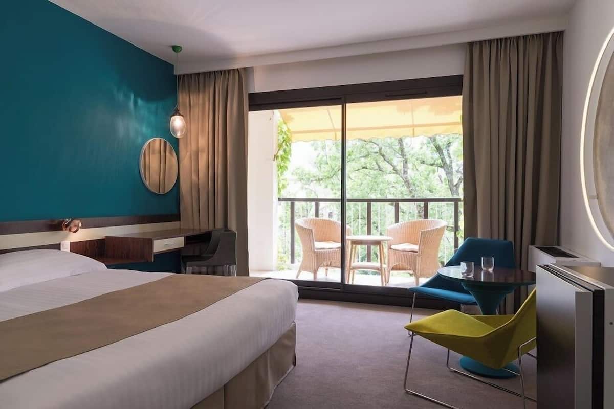 A hotel room with a bed, a desk, a chair, a table, and a balcony with chairs.