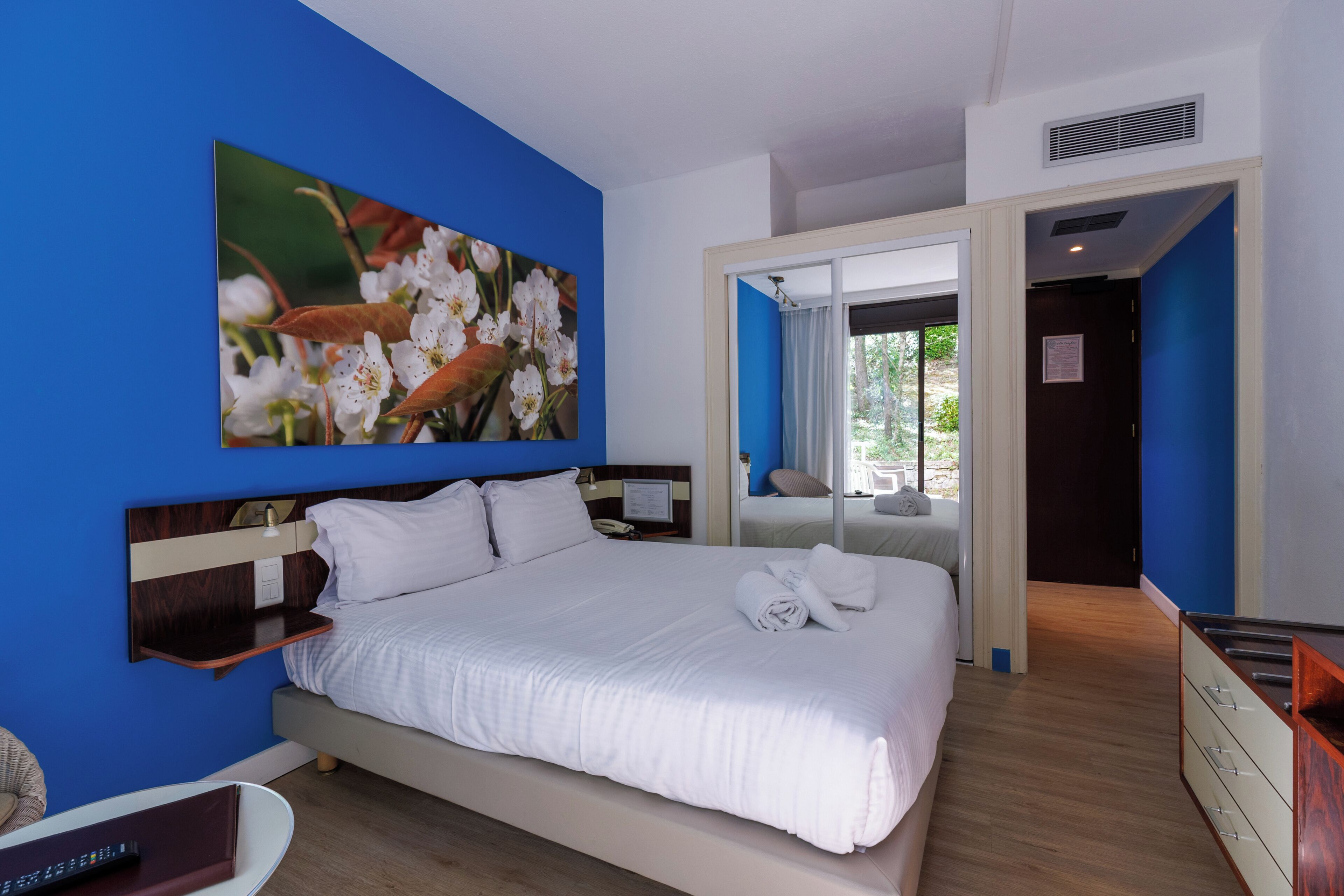 A hotel room with a bed, a nightstand, a desk, and a large window.