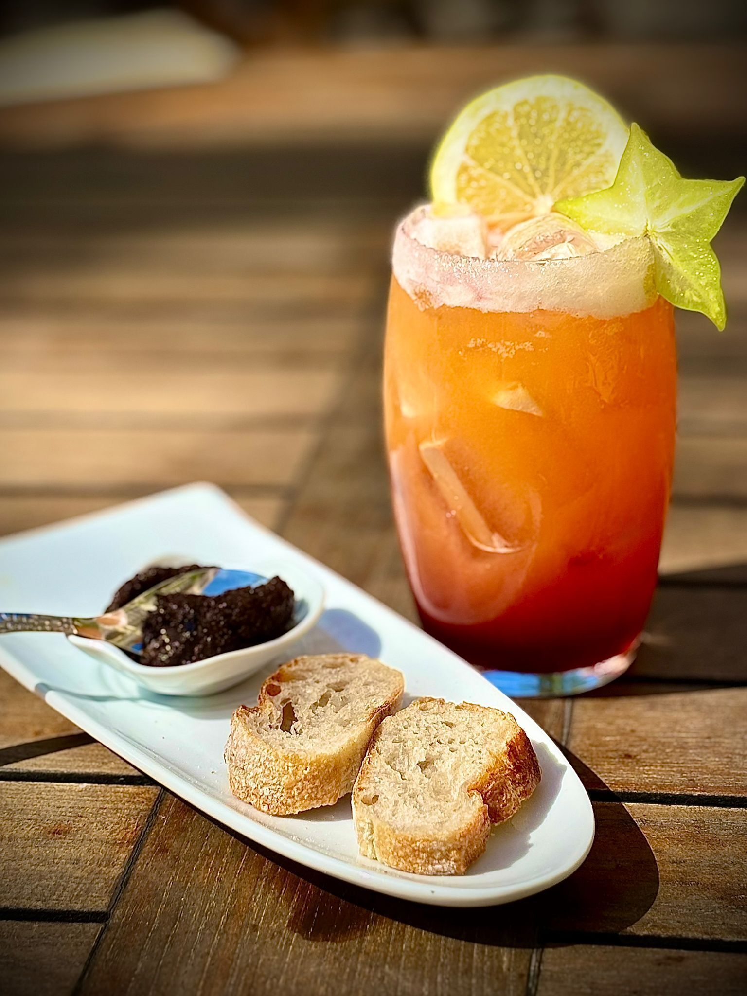A glass of iced beverage with a slice of lime on the rim, accompanied by a small bowl of chocolate and two slices of bread on a white plate.