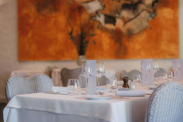 A dining table set with white tablecloth, glasses, and cutlery, with a painting in the background.