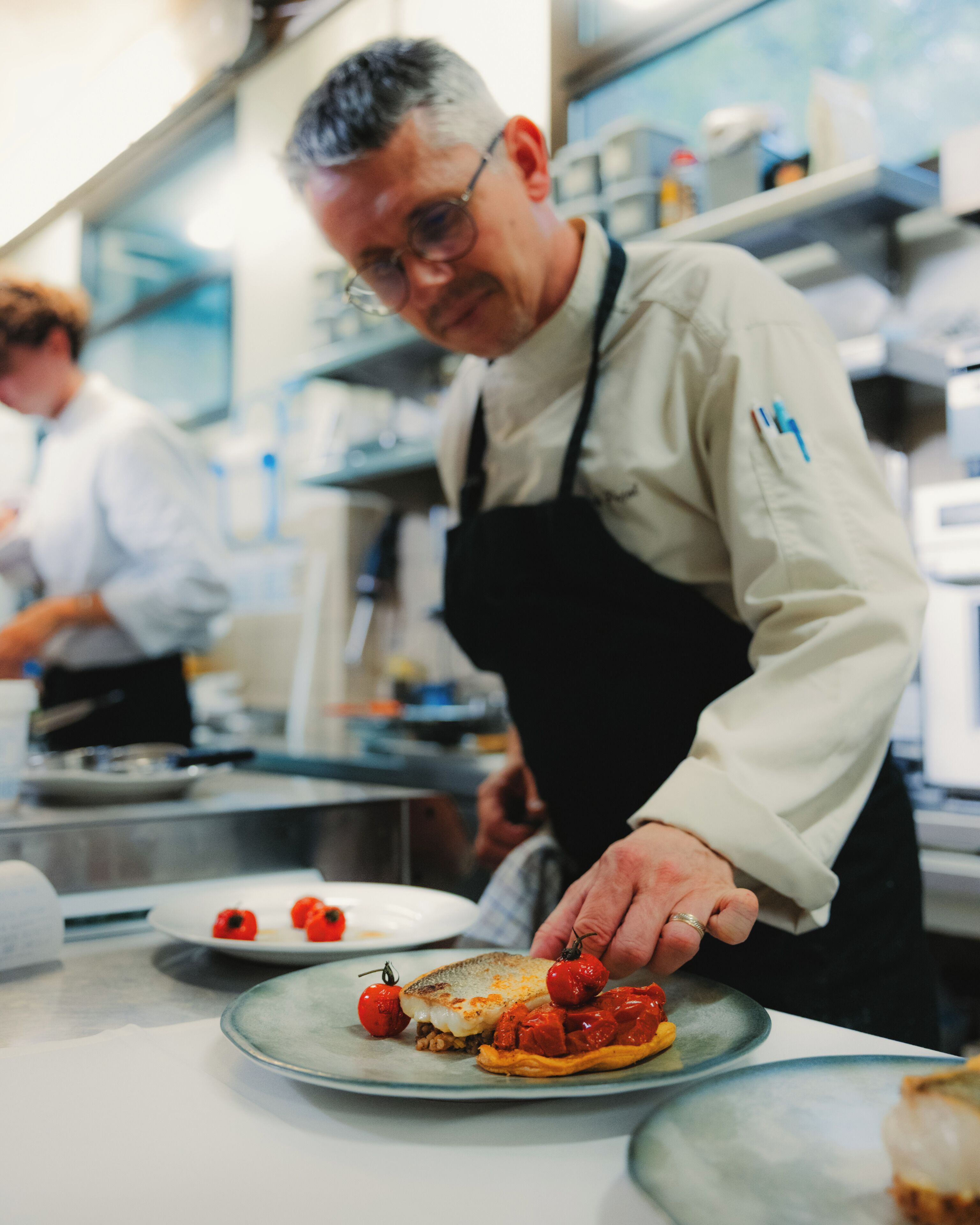 A chef in a professional kitchen with a focus on a dish with tomatoes and cheese.