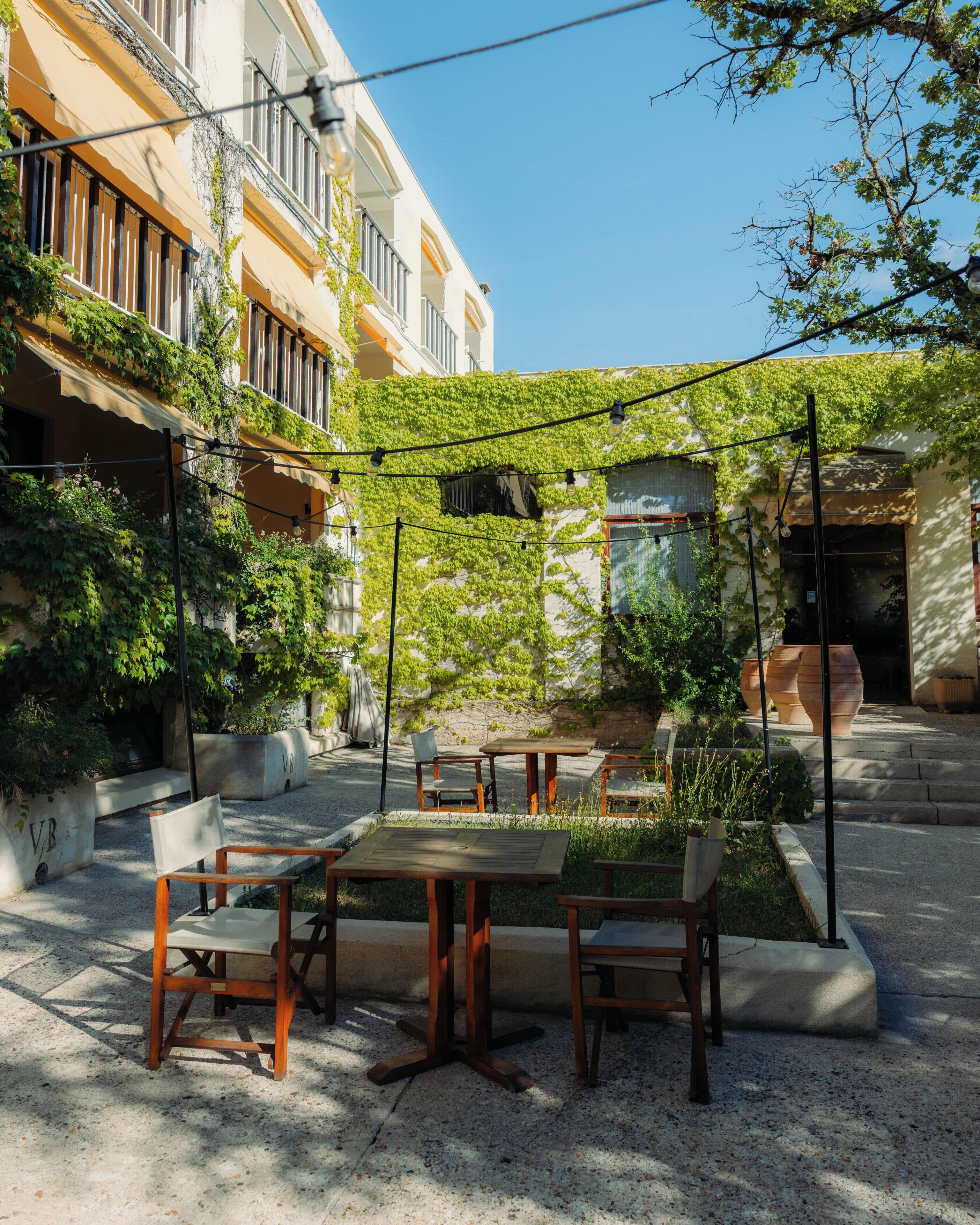 An outdoor seating area with tables and chairs, surrounded by greenery and a building with a white facade.