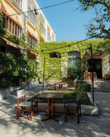 Un área de estar al aire libre con mesas y sillas, rodeada de follaje y un edificio de fachada blanca.