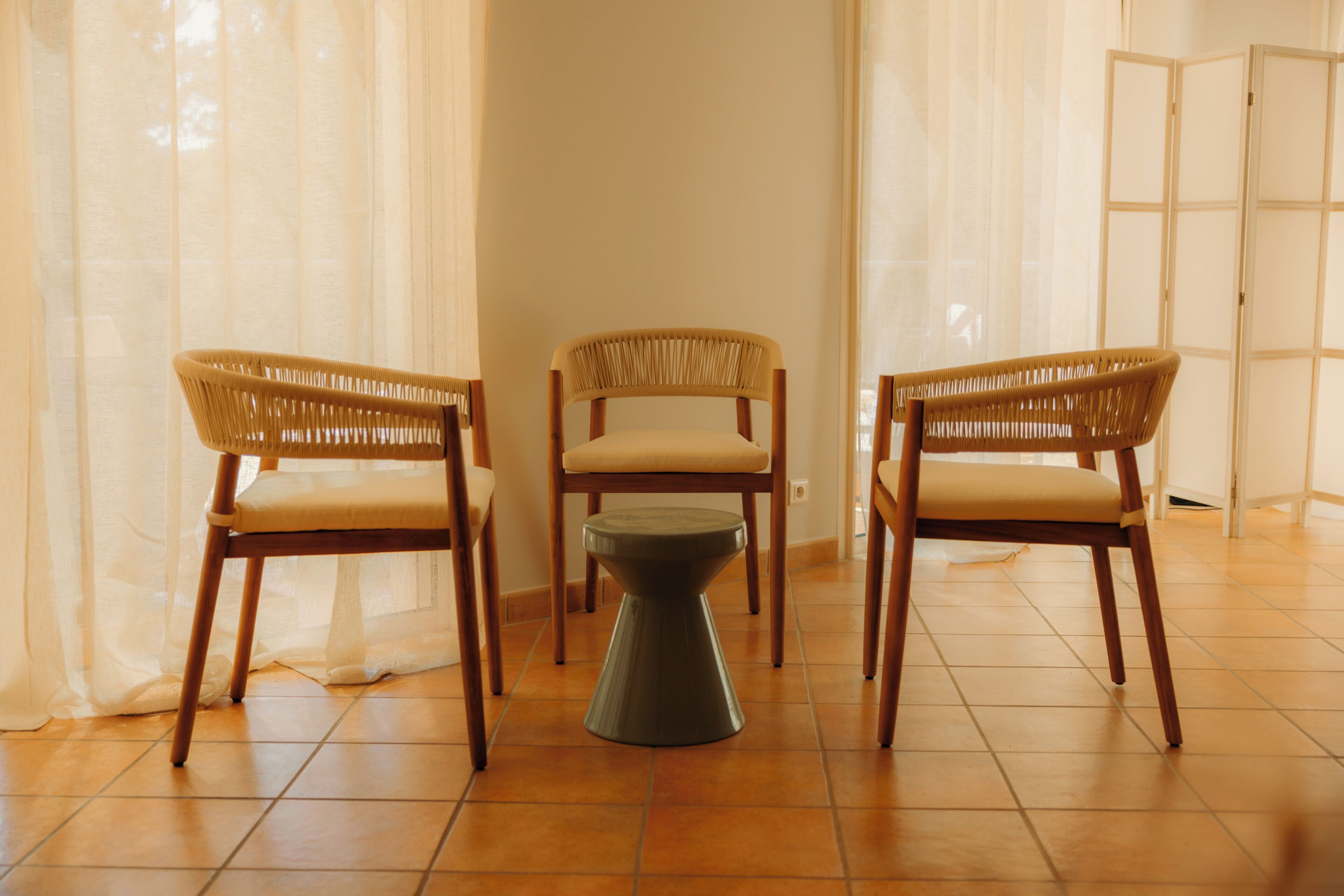 Una habitación con tres sillas de mimbre y una pequeña mesa, suelo de baldosas y cortinas de encaje.