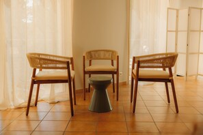 Una habitación con tres sillas de mimbre y una pequeña mesa, suelo de baldosas y cortinas de encaje.