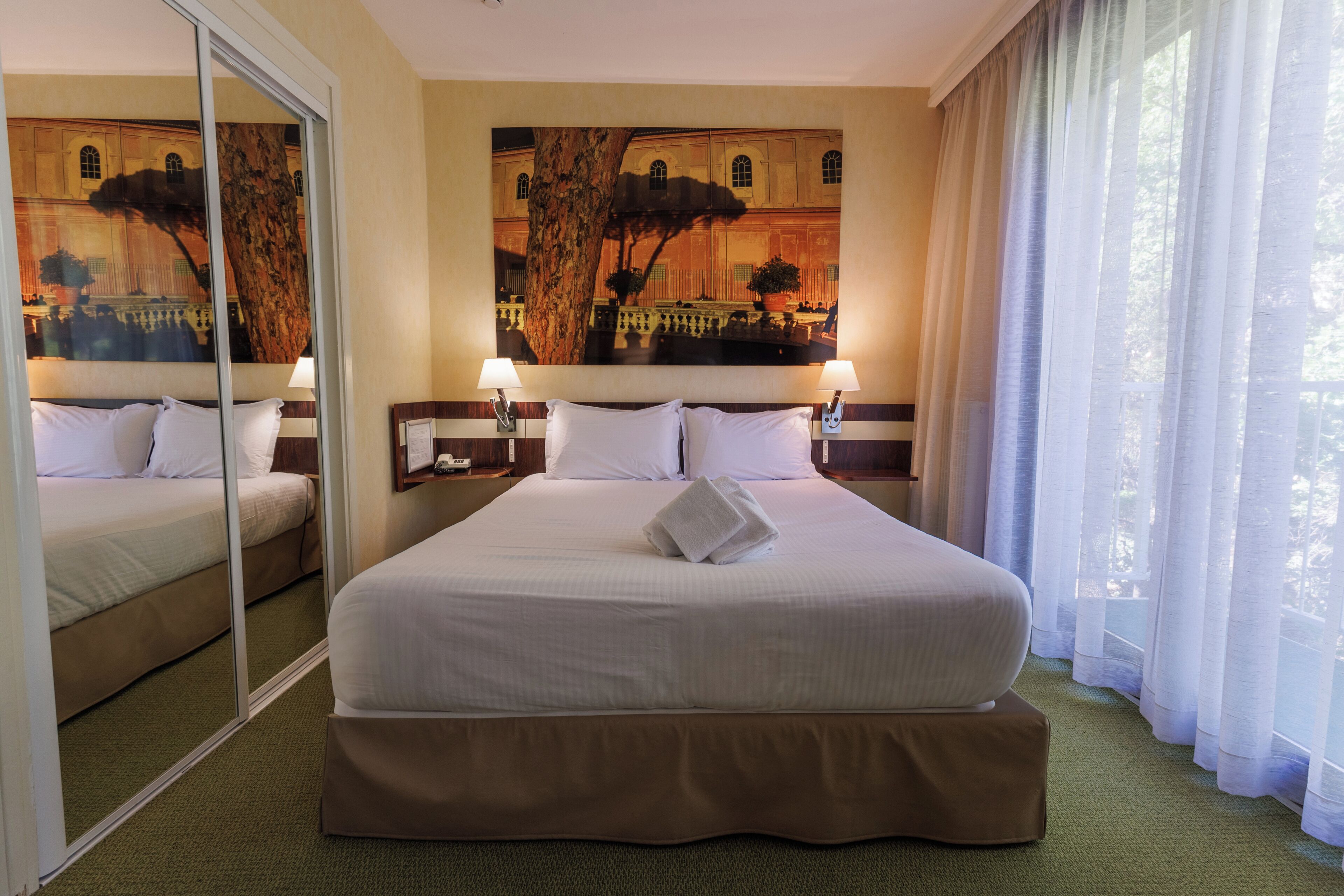 Habitación de hotel con una cama grande, dos mesitas de noche con lámparas, un espejo amplio y un cuadro en la pared.