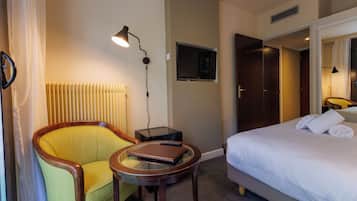 Habitación de hotel con cama, una silla, una mesita y un radiador. Hay un televisor montado en la pared.