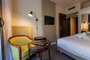 A hotel room with a bed, a chair, a small table, a TV mounted on the wall, and a radiator.