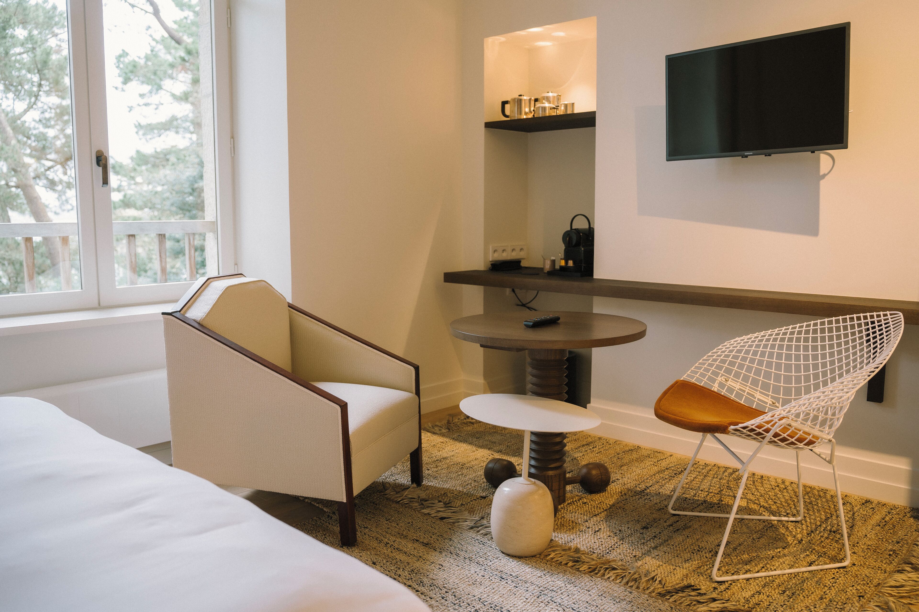 Et moderne hotelværelse med en seng, to stole, et rundt bord, et fladskærms-tv på væggen og et vindue med udsigt til træer.
