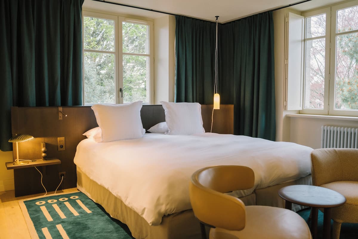 A hotel room with a bed, a chair, a small table, a lamp, and a view of trees through the window.