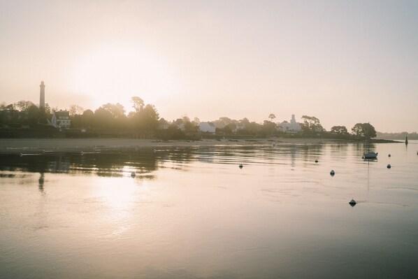 A calm river with boats, a distant lighthouse, and buildings along the shore.