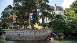 A stone wall with flags, a building with a sign reading "A TRI MEN", and a body of water. - Villa Tri Men (Combrit)
