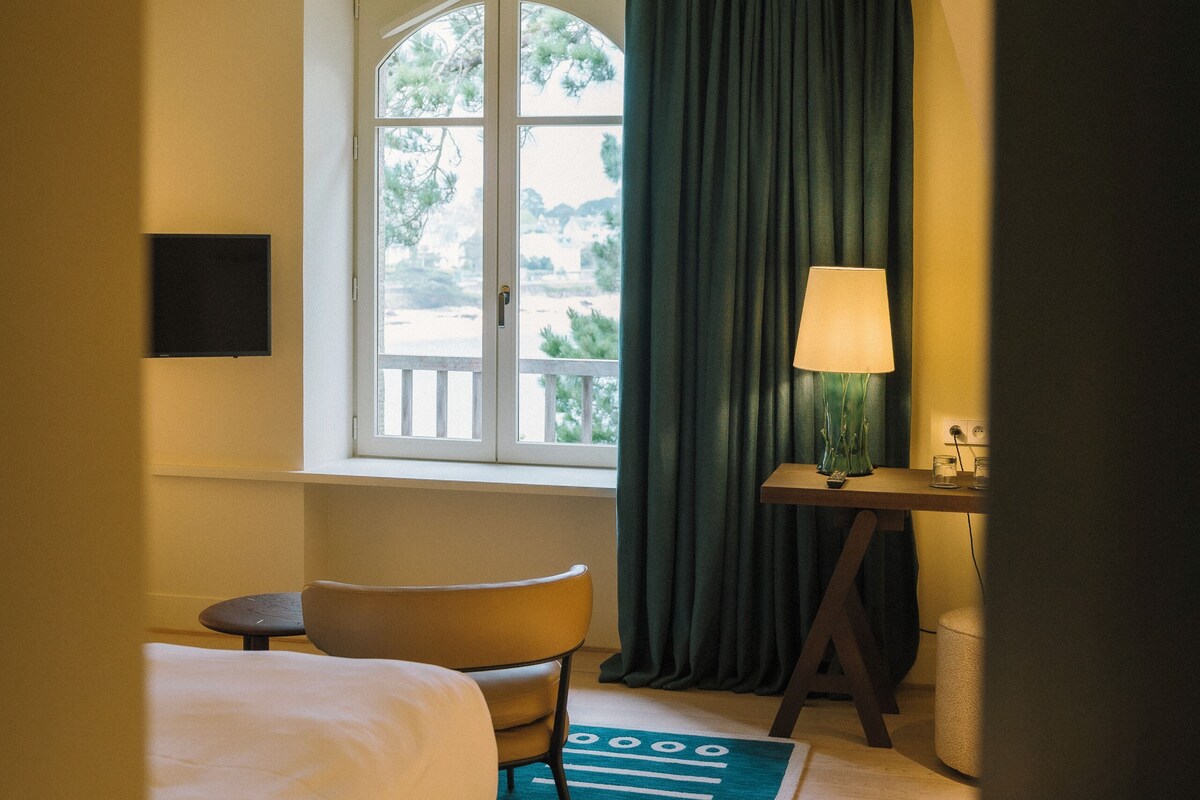 A hotel room with a bed, a chair, a desk with a lamp, a window with curtains, and a view of the outdoors.