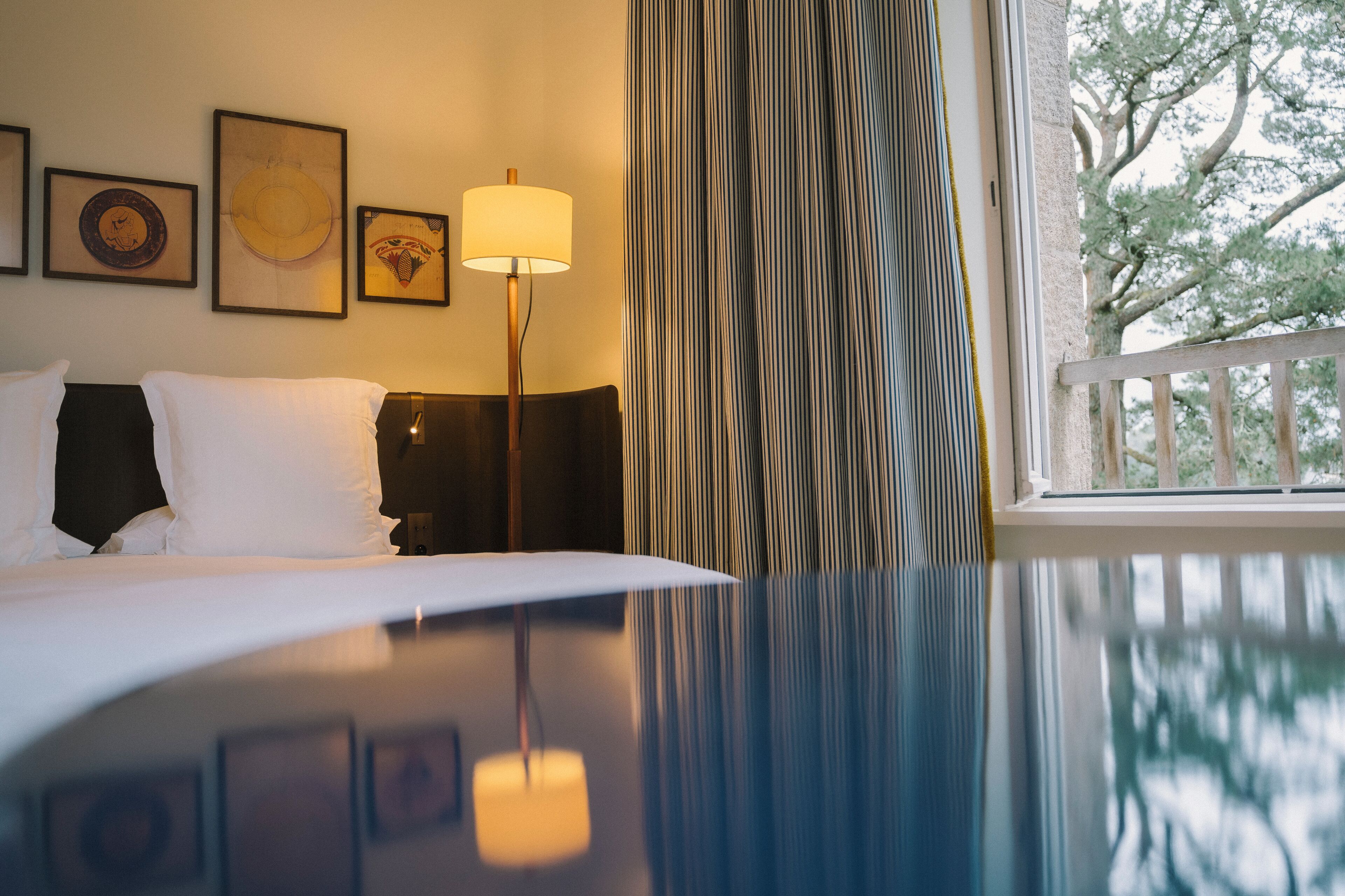 Una habitación de hotel con una cama, una lámpara, un ventanal con cortinas y cuadros en la pared.