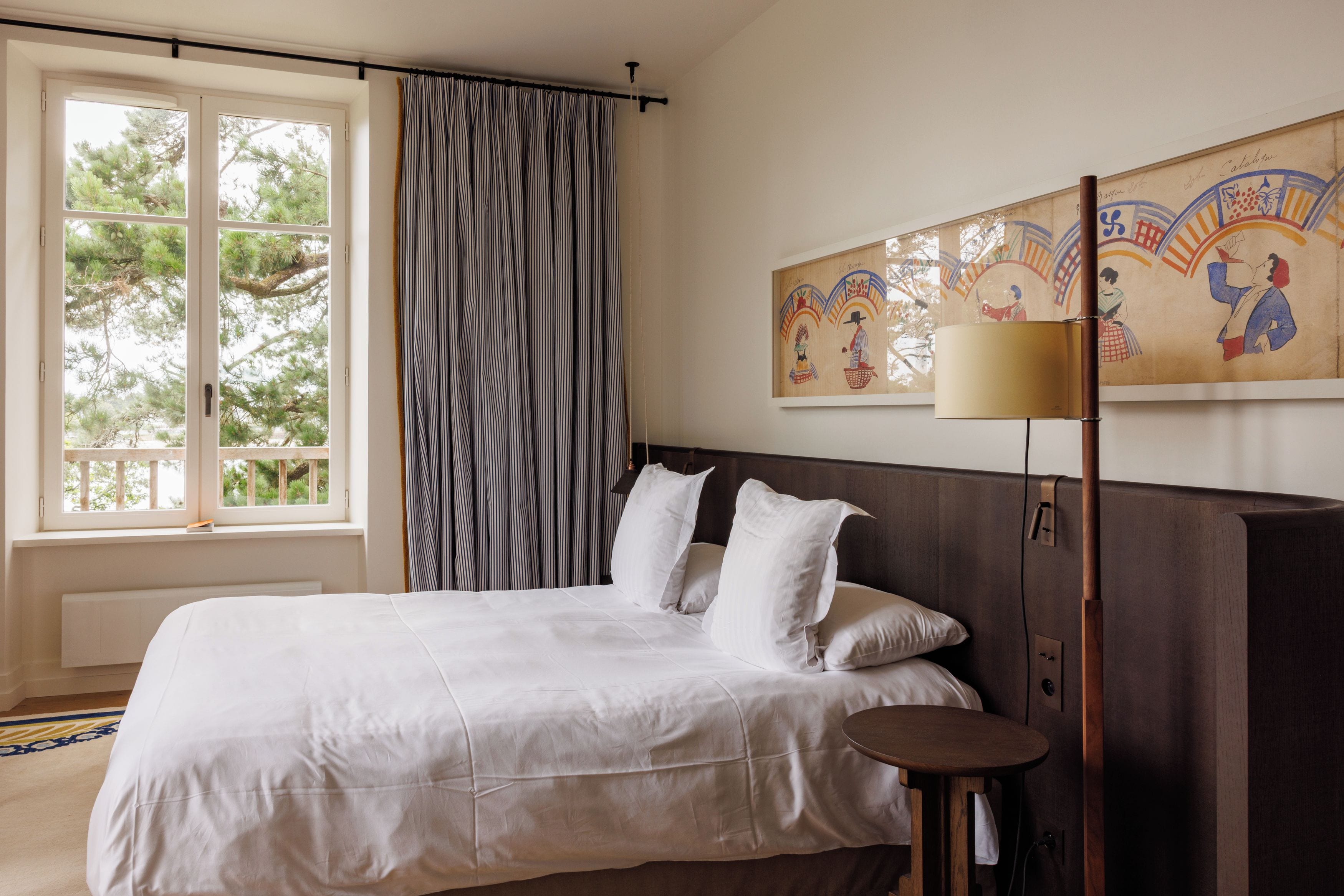 A bedroom with a large bed, a bedside table, a lamp, and a window with curtains.