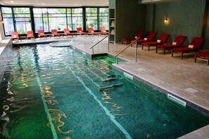 Une piscine intérieure à l’eau claire, entourée de chaises longues rouges et bordée d’une allée carrelée.