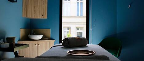 Une pièce avec un meuble en bois, un bol posé sur un comptoir, une chaise verte et une fenêtre donnant sur des immeubles.