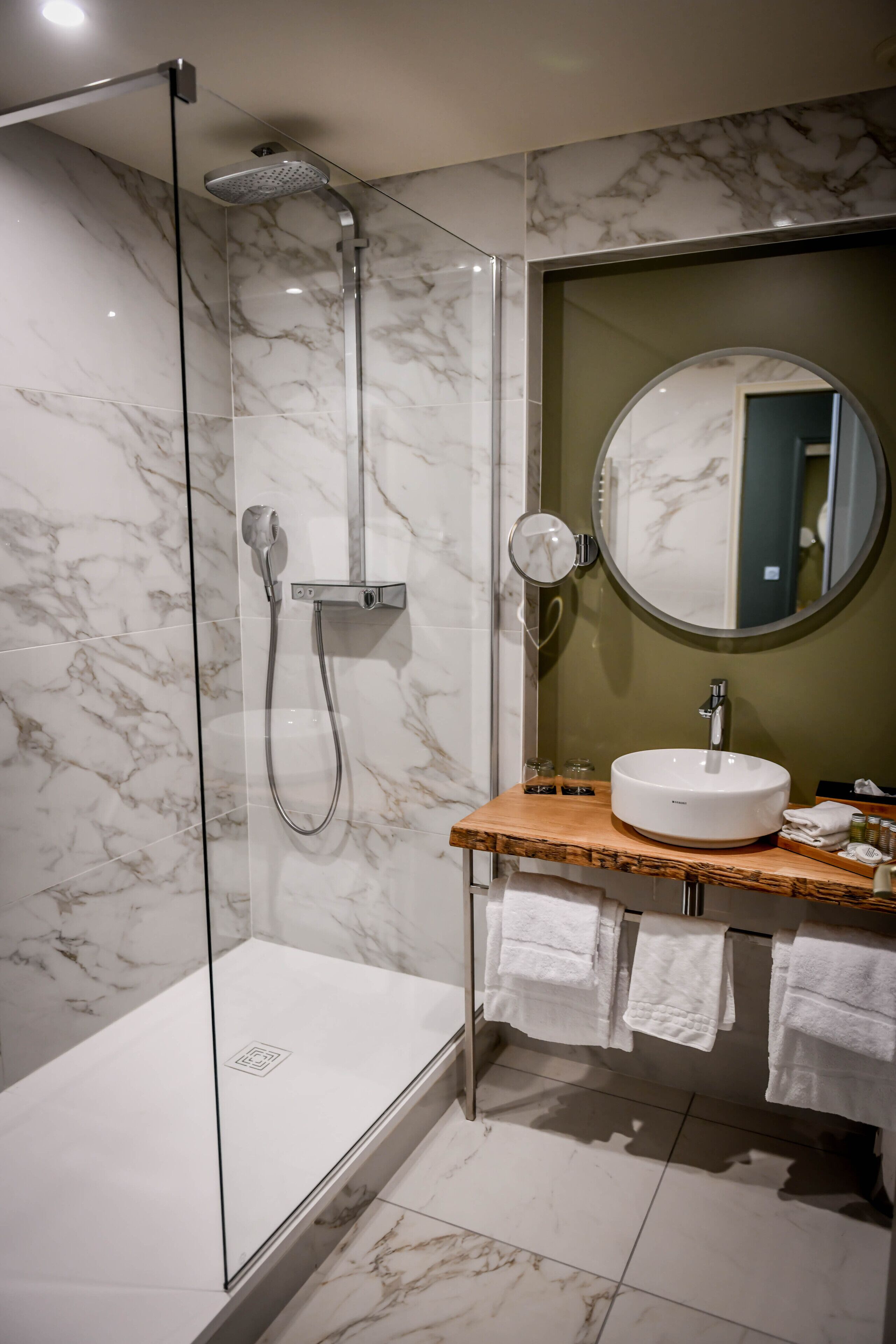 A modern bathroom with a glass-enclosed shower, a marble-tiled floor, a wooden vanity with a sink, and a round mirror.
