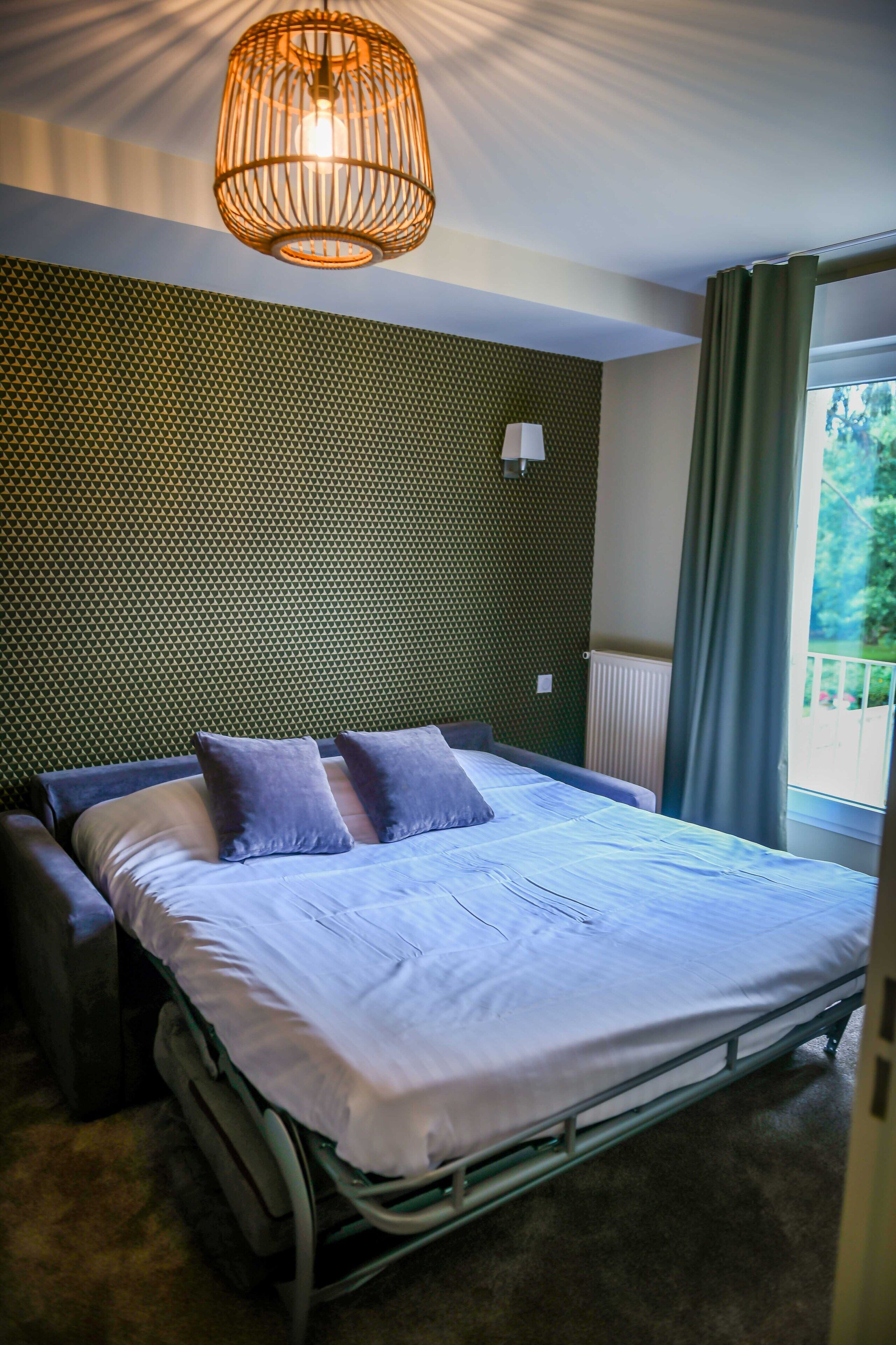 Quarto com cama, mesa de cabeceira, luminária pendente, parede estampada e janela com cortinas.